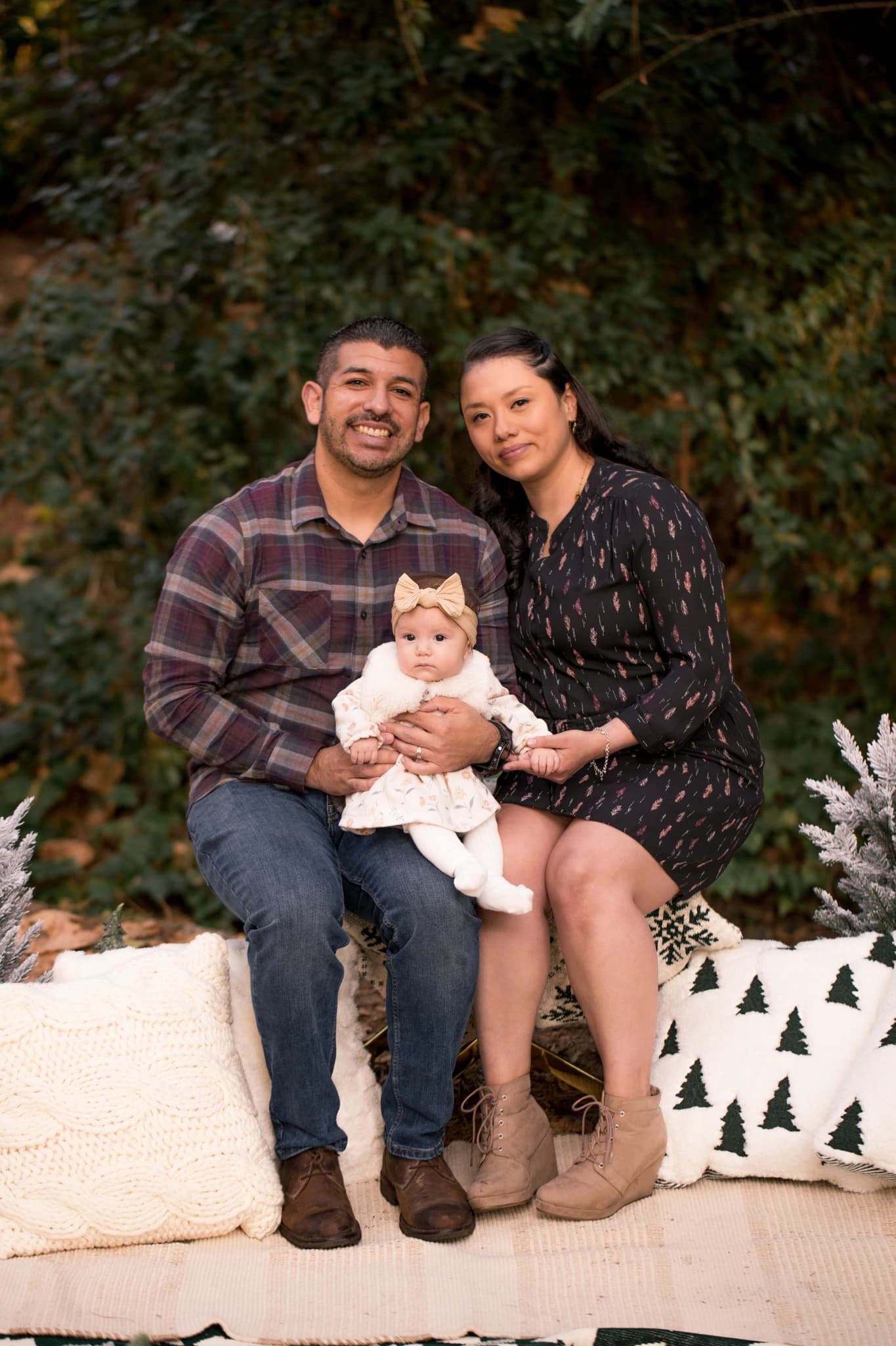 Parents holding their baby on a cream blanket beside frosted trees and Christmas cushions